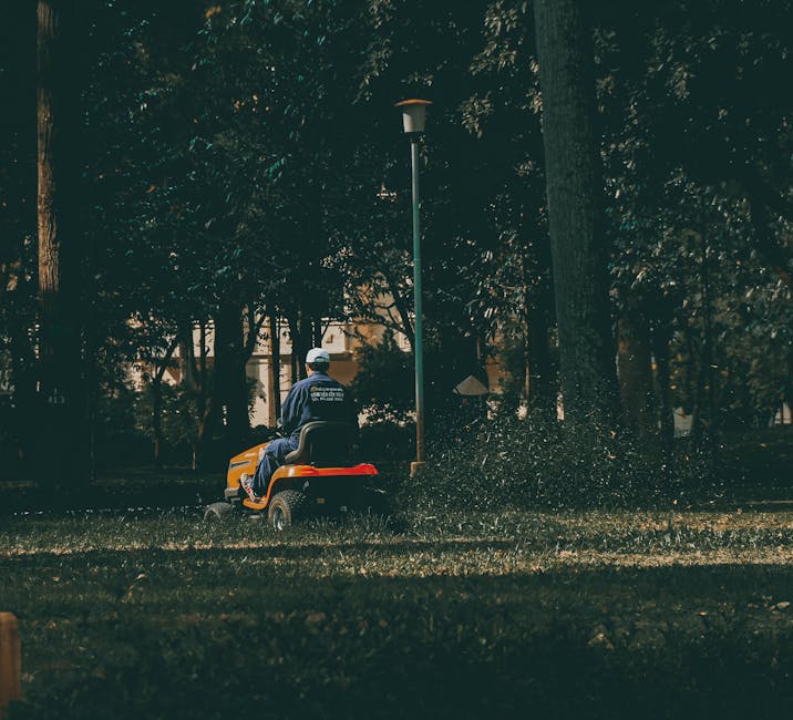 A grounds maintenance worker operating a ride-on lawn mower in a lush, green park or garden area during daylight hours. The scene features well-maintained grass with visible mowing patterns, bordered